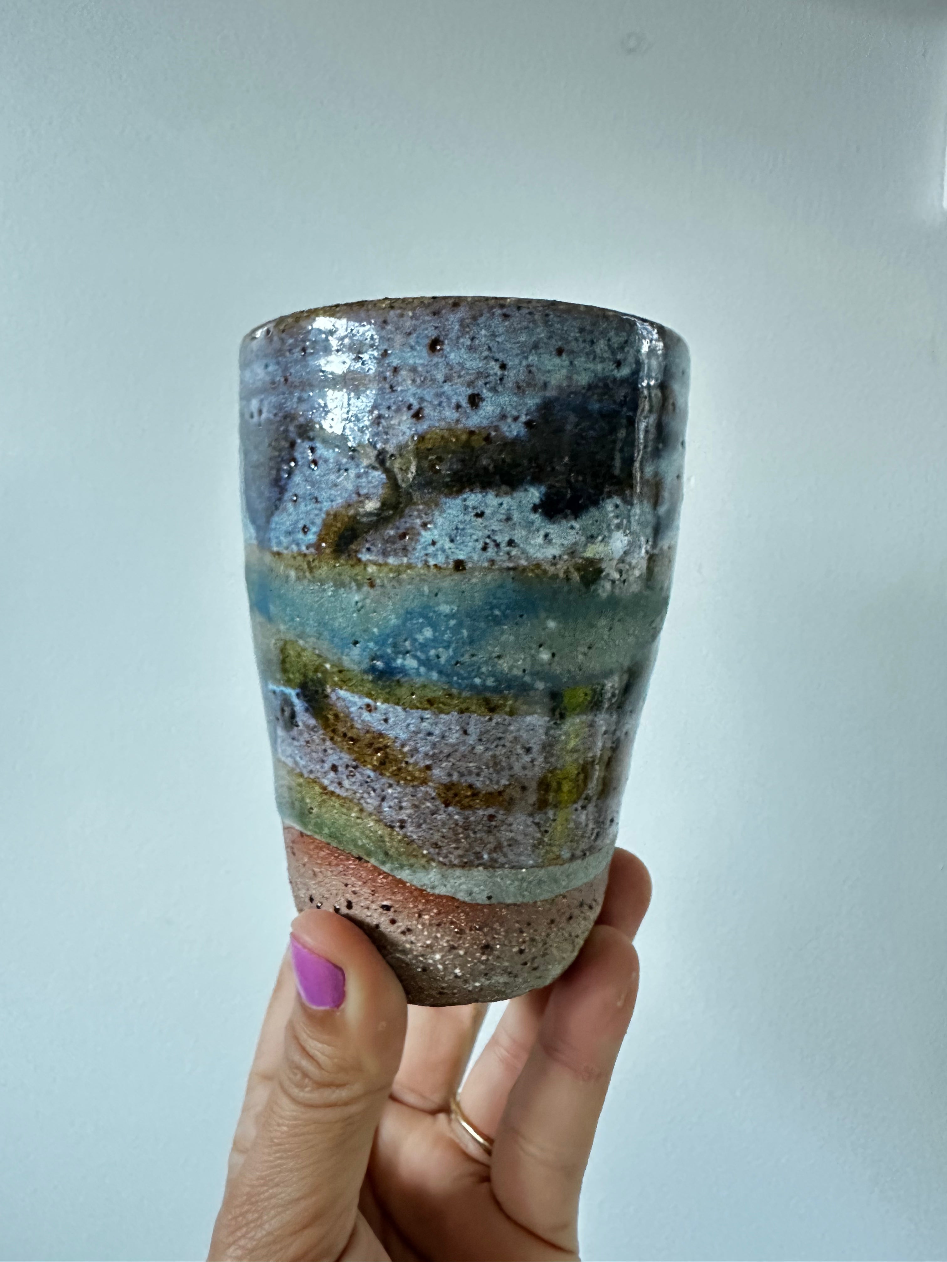 Hand holding a ceramic cup with multicolored glaze against a plain background