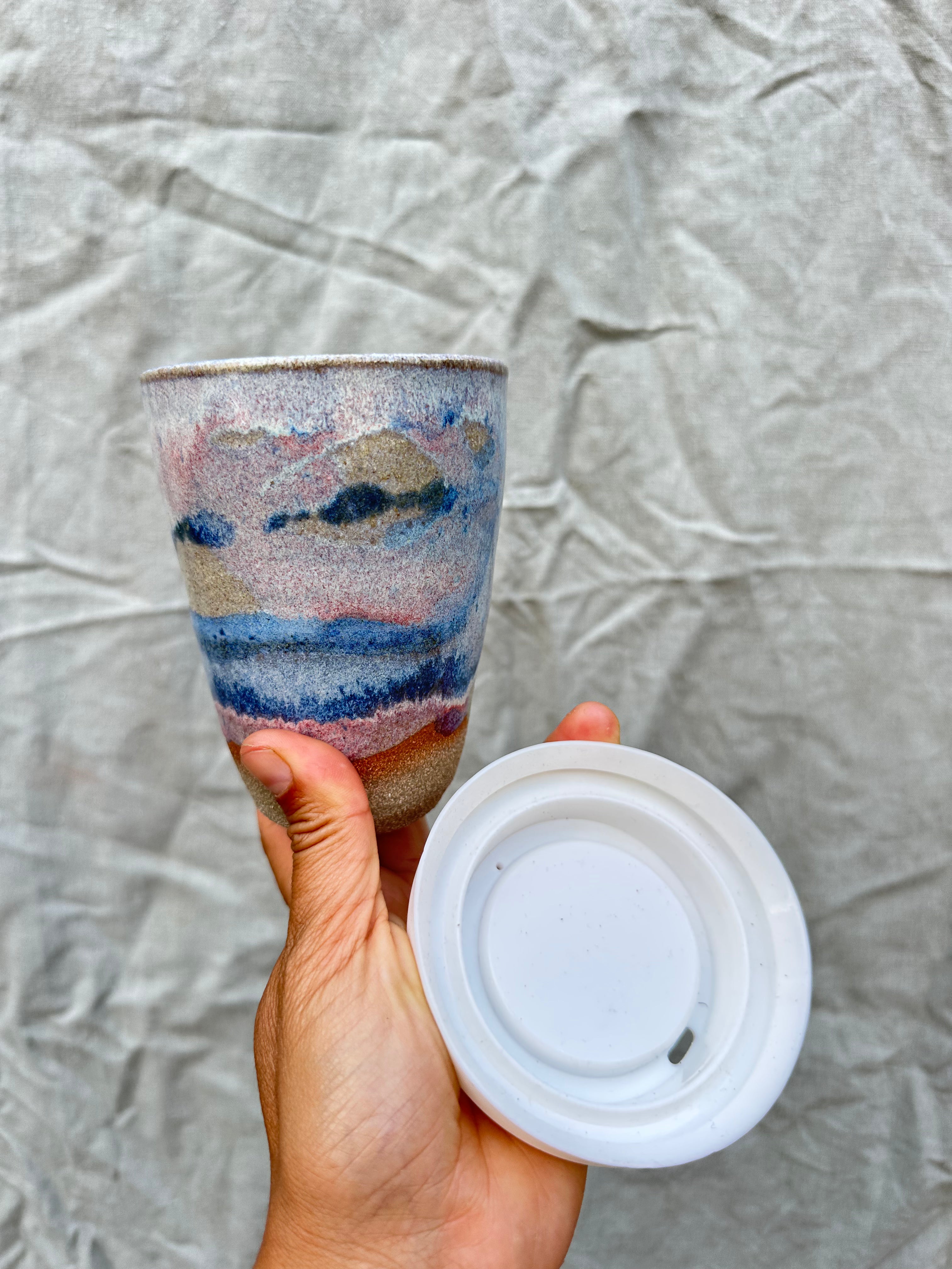 Hand holding a ceramic tumbler with a colorful design and a silicone lid against a textured gray background