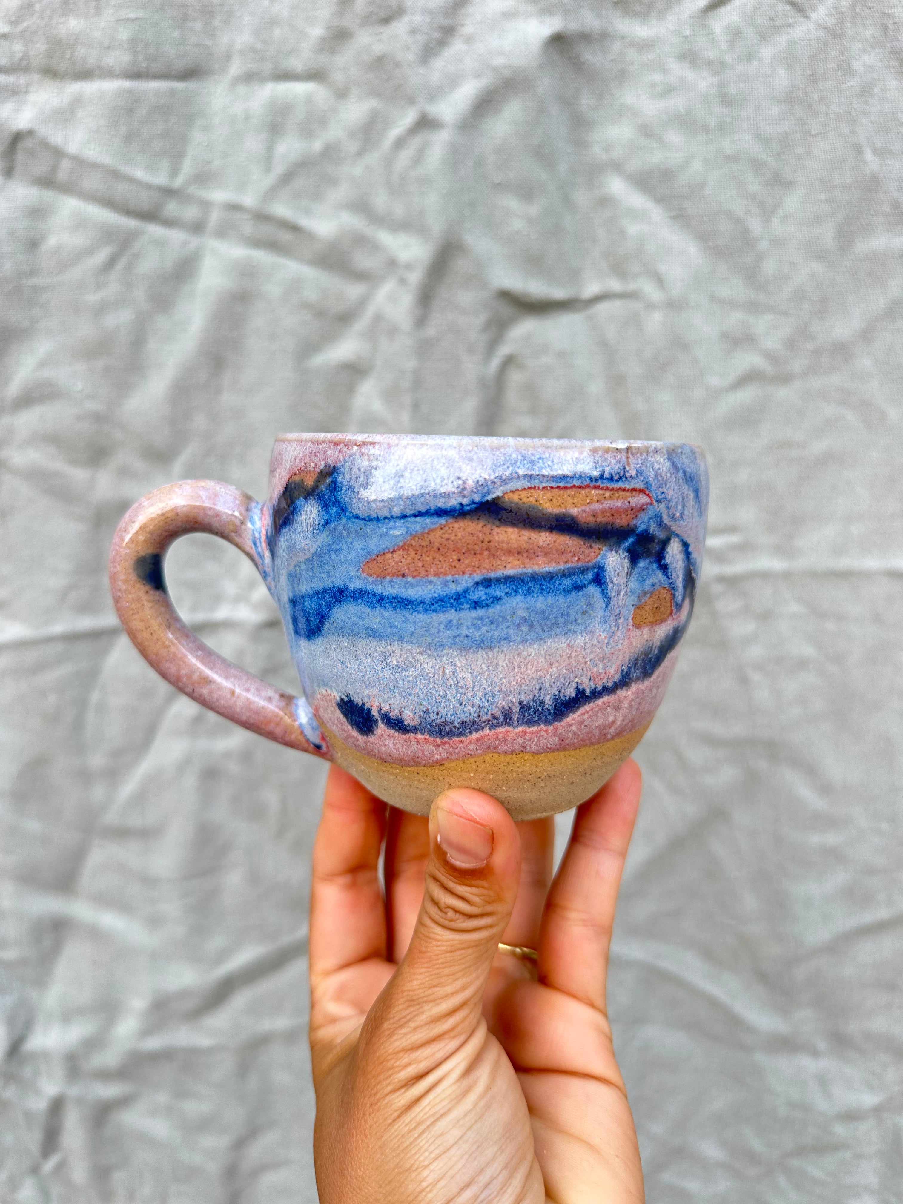 Hand holding a ceramic mug with blue and brown abstract design against a textured gray background