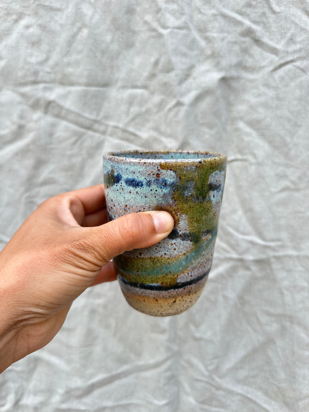 Hand holding a ceramic cup with multicolored glaze against a textured grey background