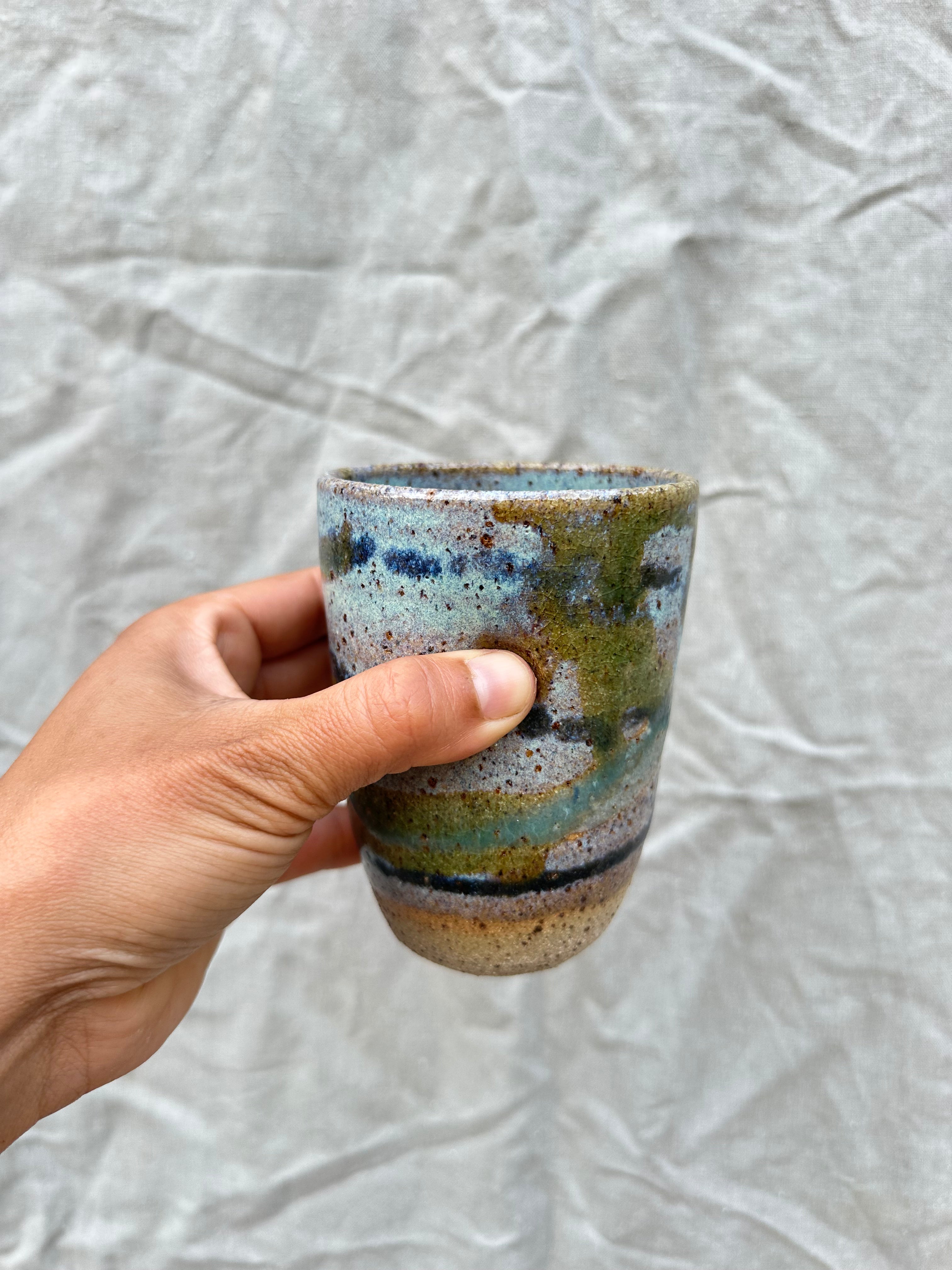 Hand holding a ceramic cup with multicolored glaze against a textured grey background