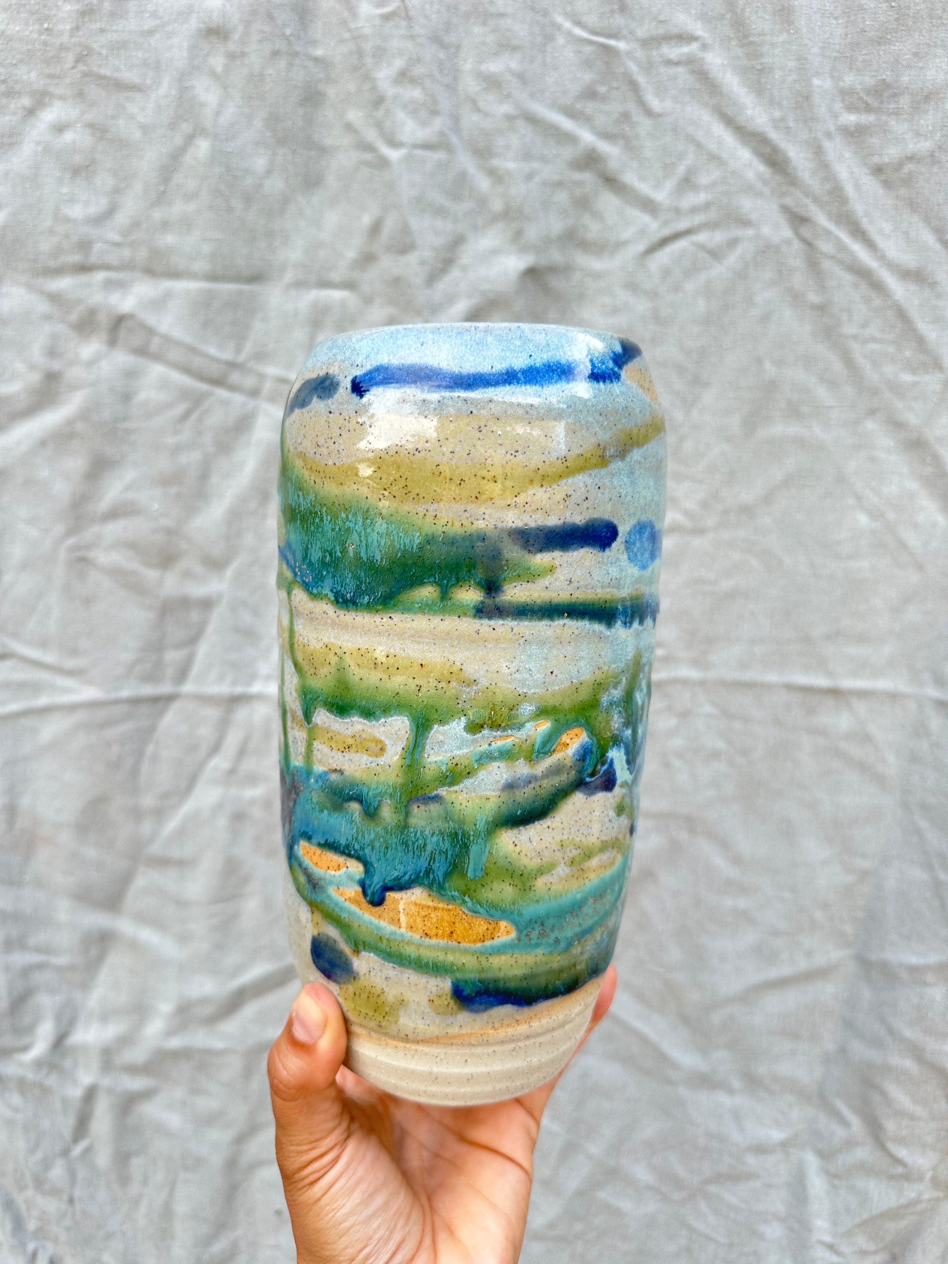 Hand holding a ceramic vase with a scenic design against a textured gray background