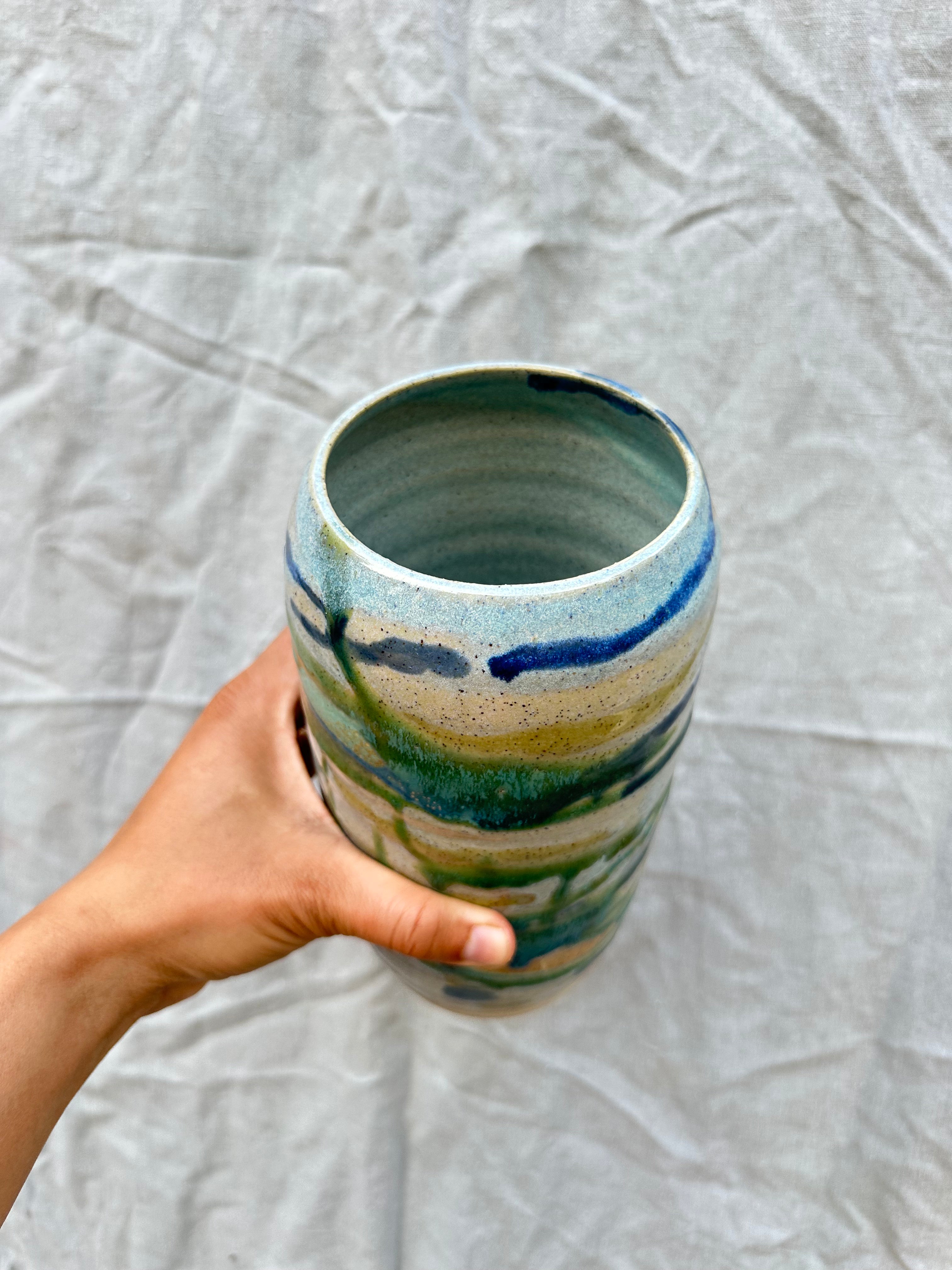 Hand holding a ceramic vase with blue and green stripes on a light gray background